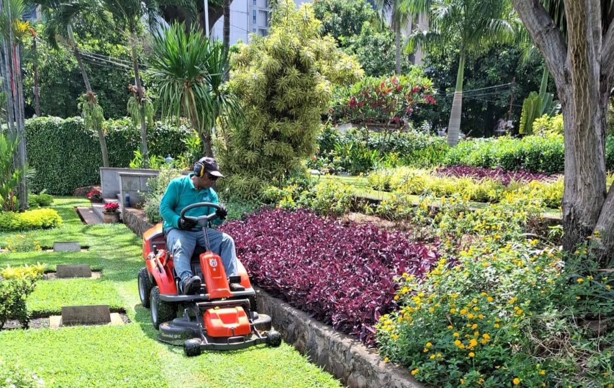 A person rides a red riding lawn mower across a grassy cemetery, with lush green grass, stone markers, and colorful flower beds.