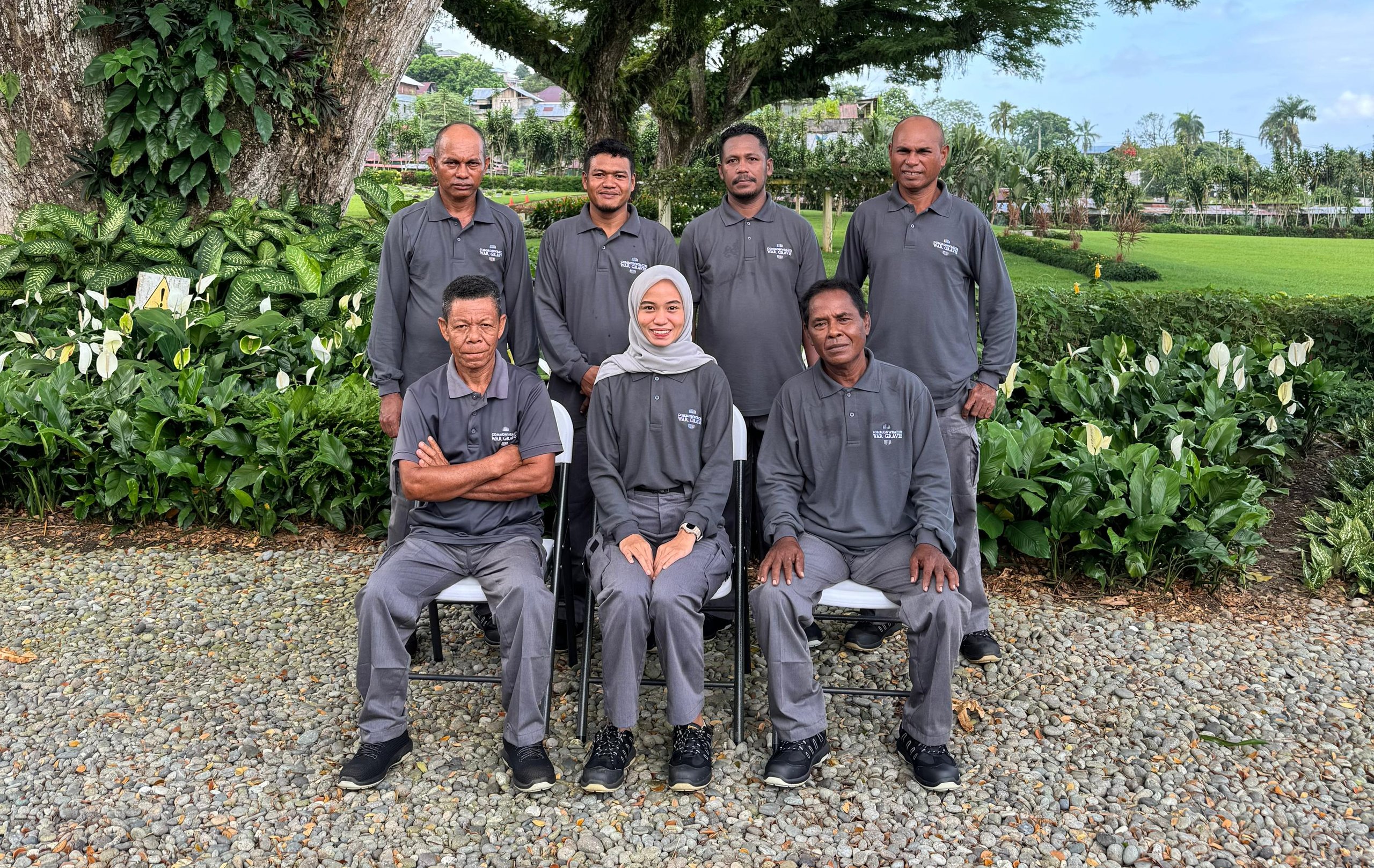 Group of eight individuals, seven men and one woman, pose for a photo outdoors. They are wearing matching grey uniforms with a logo. The group is arranged in two rows, with three people seated in front and five standing behind them. They are positioned in front of a large, old tree covered in vines and ferns, with a lush green lawn and some buildings visible in the background.