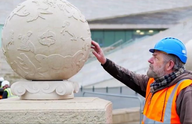 A man in a hard hat and safety vest gently touches a large, carved stone sphere depicting various figures and animals.