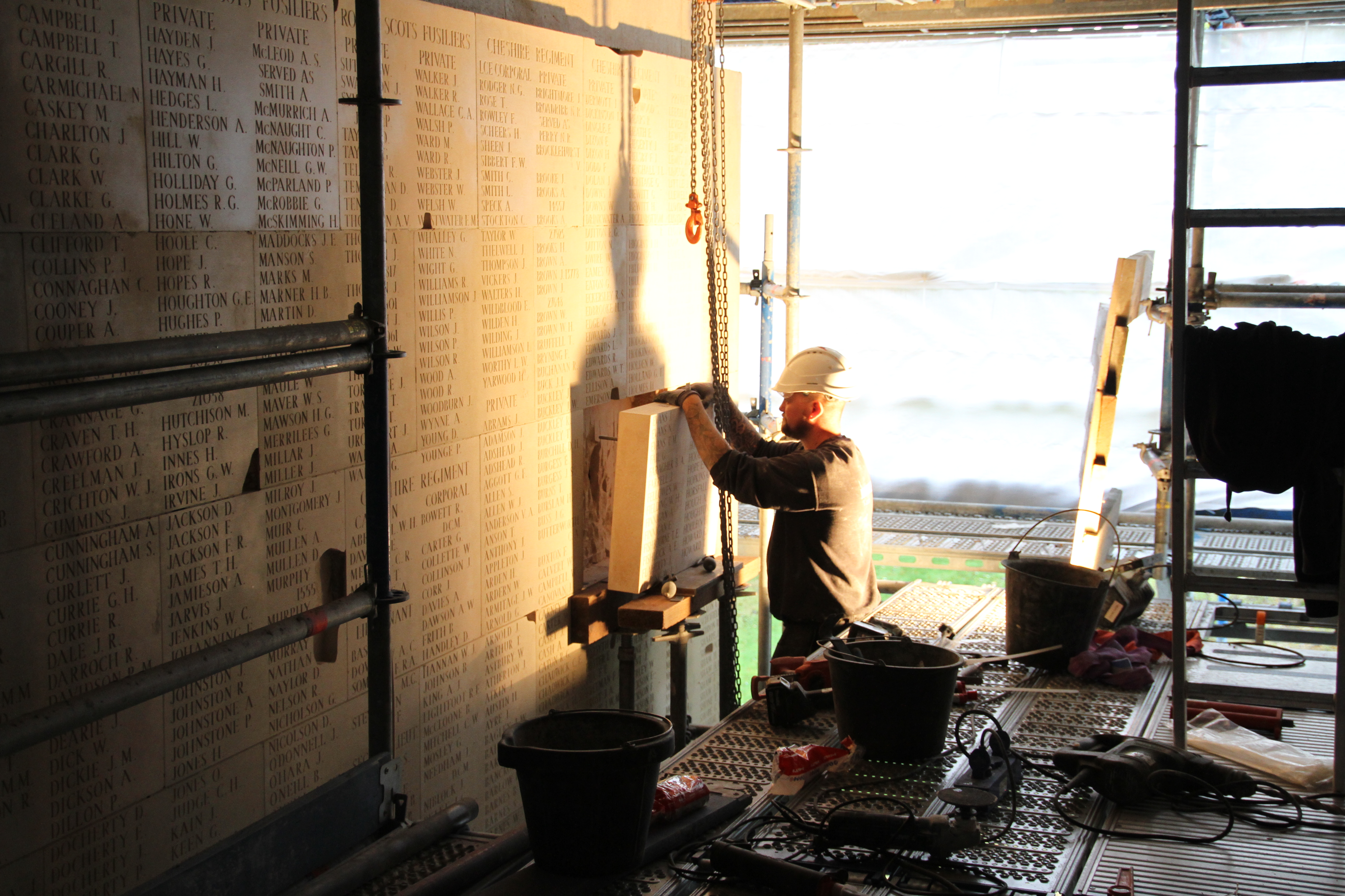 Worker in hard hat adjusts a chain near a stone memorial wall listing names. Tools and buckets are scattered nearby.