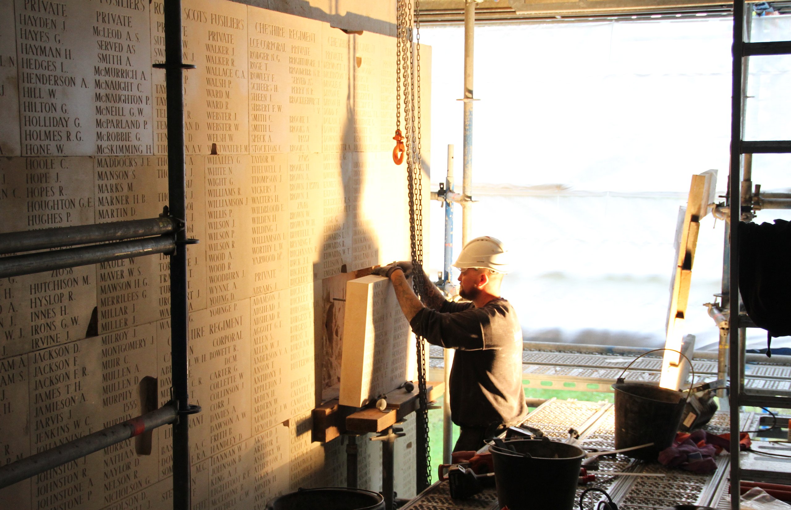 Worker in hard hat adjusts a chain near a stone memorial wall listing names. Tools and buckets are scattered nearby.