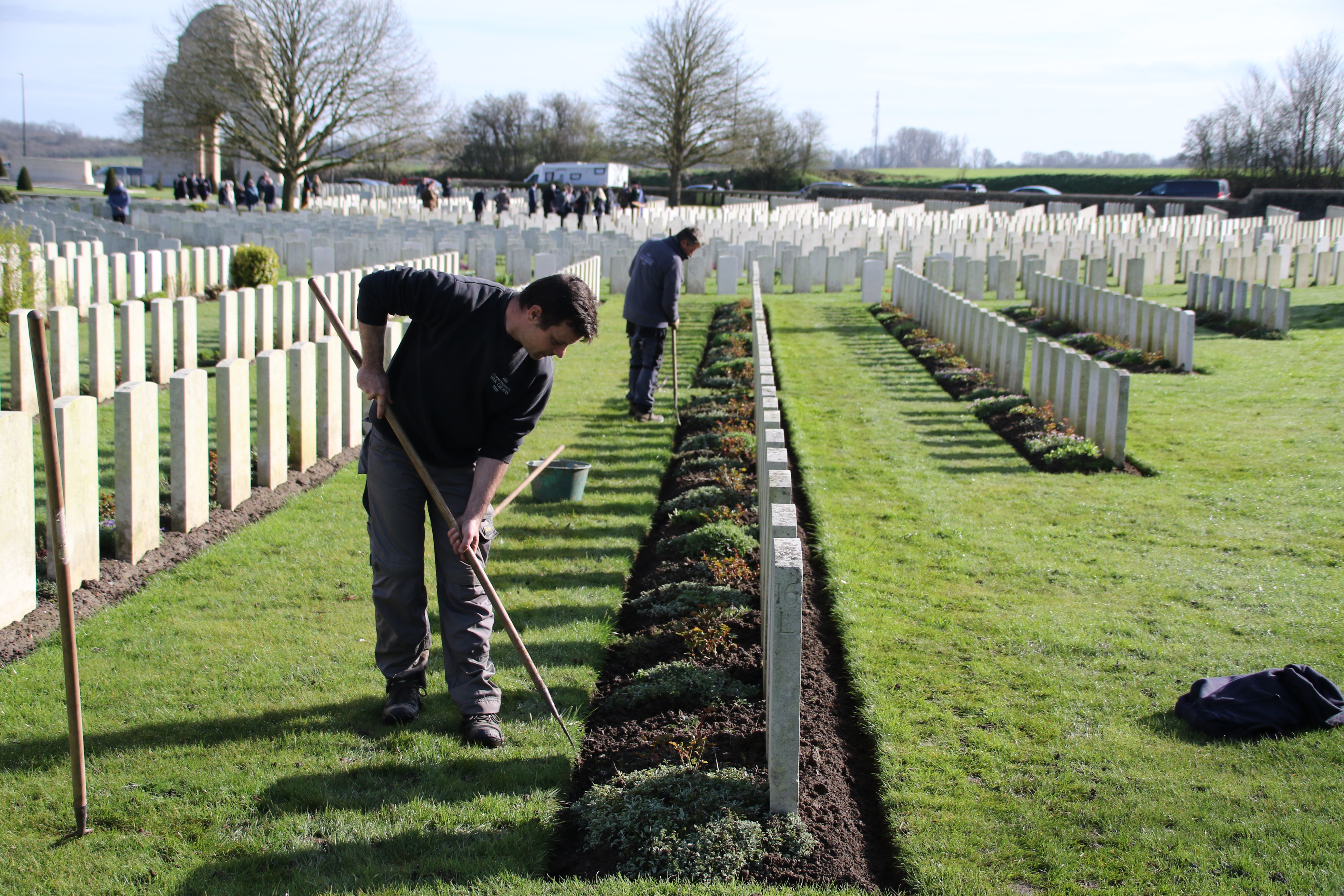 Workers tending graves in a cemetery. Rows of white headstones stretch into the distance under a clear sky.