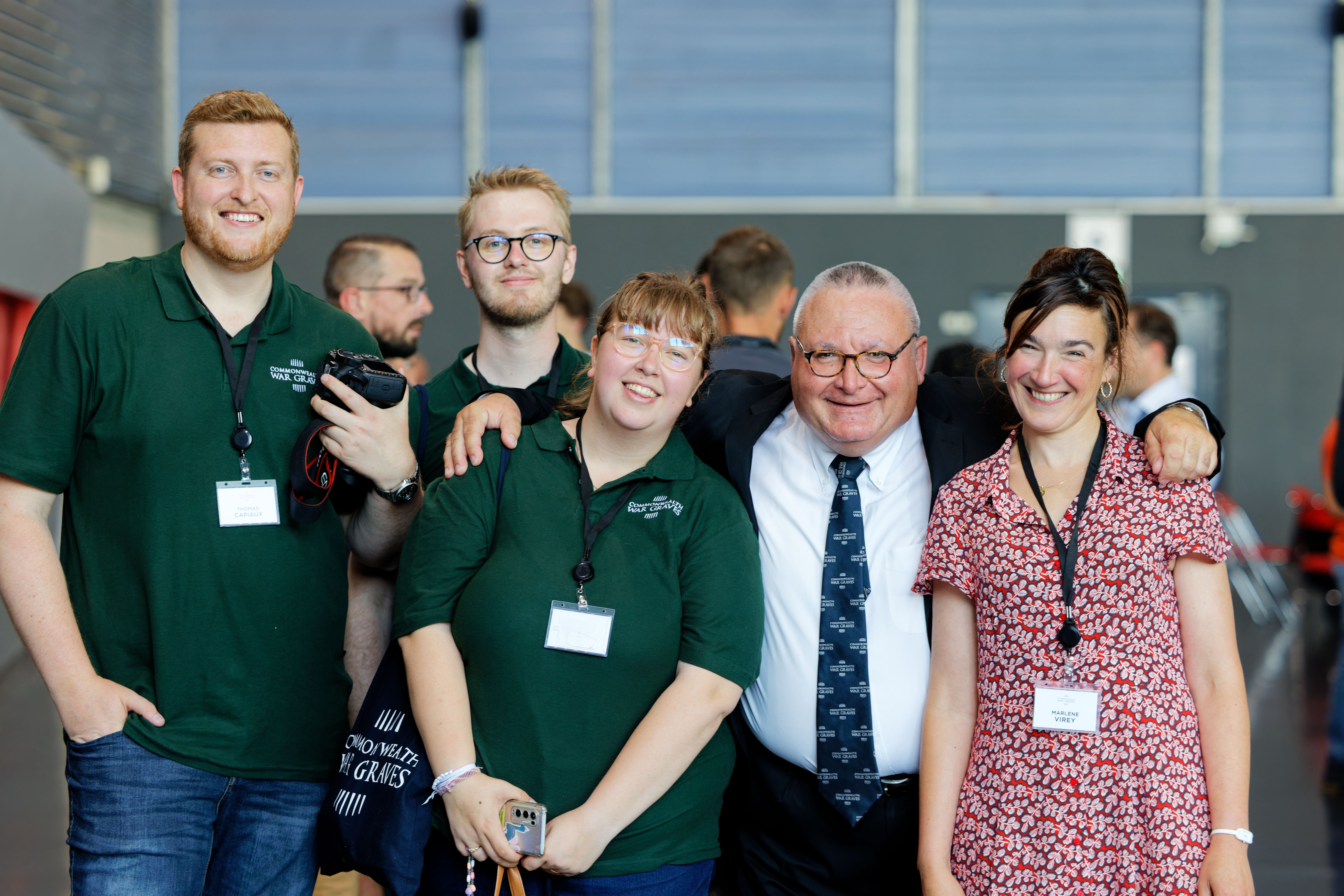 A group of five people, three wearing green polo shirts with "Commonwealth War Graves" badges, smile for a photo. A man in a suit and tie is in the center.
