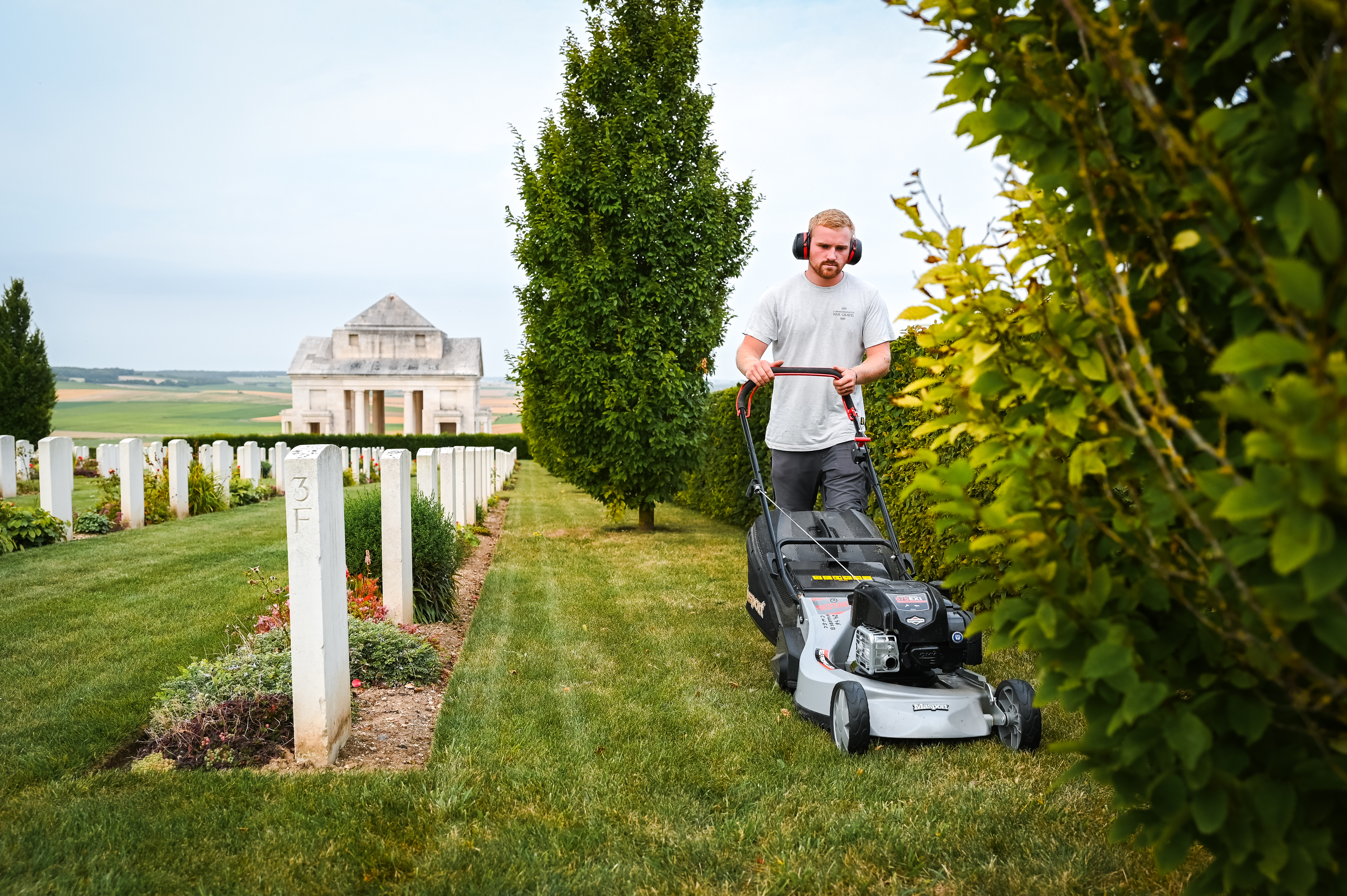 A man in headphones mows the lawn in a cemetery with rows of white headstones and a large building in the background.