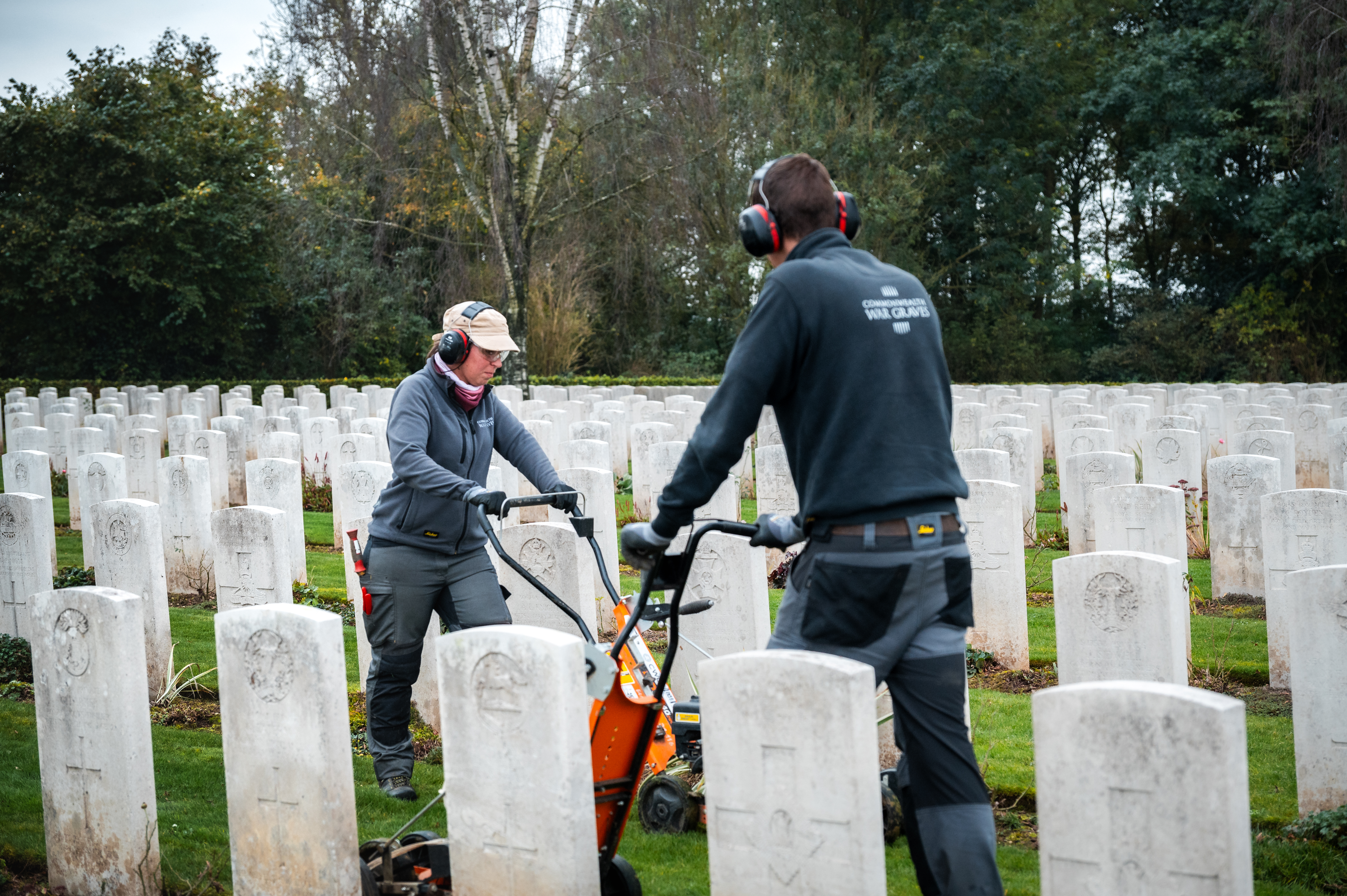 Two people, one a woman with a cap and ear protection, are operating a lawn mower between rows of white headstones in a cemetery.