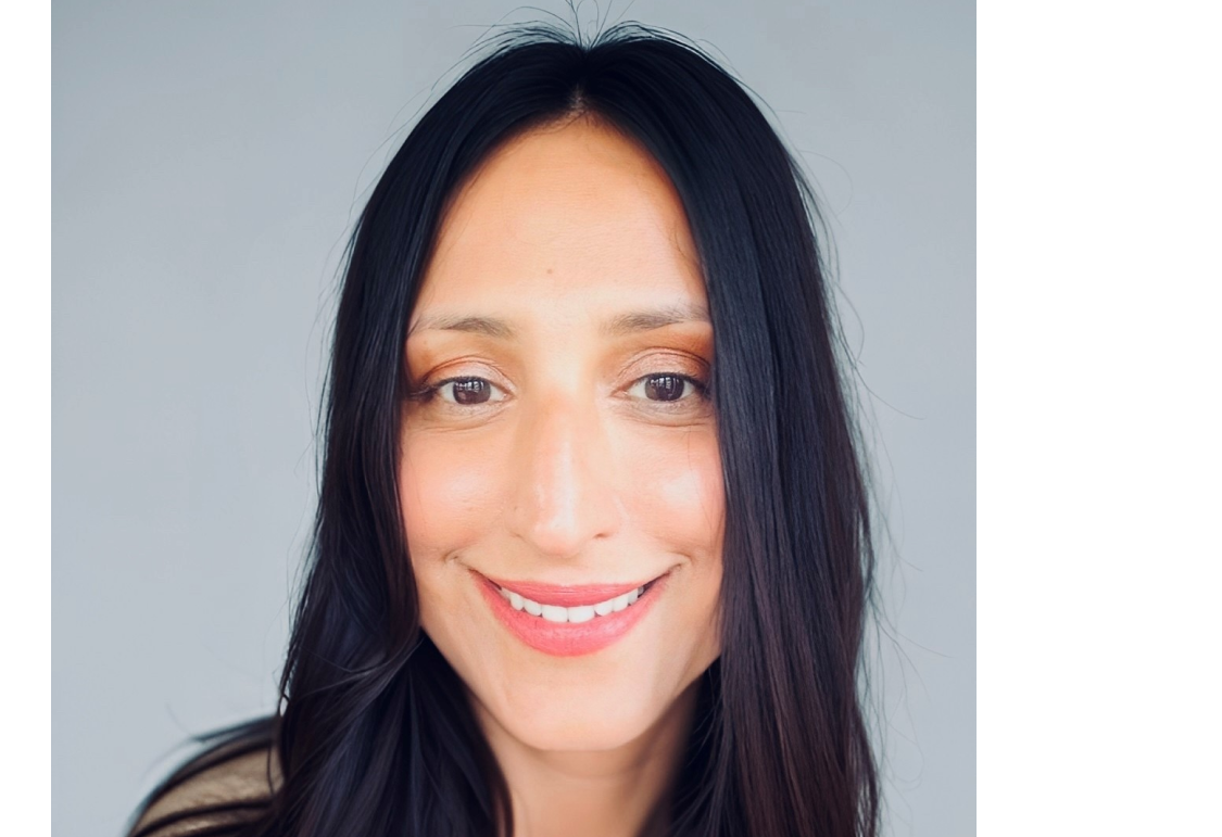 A smiling woman with long, dark hair and brown eyes, wearing a light-colored sweater against a gray background.