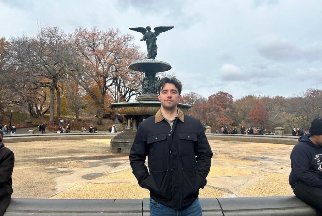 A man stands in front of Bethesda Fountain in Central Park on a cloudy day, with autumn foliage in the background.