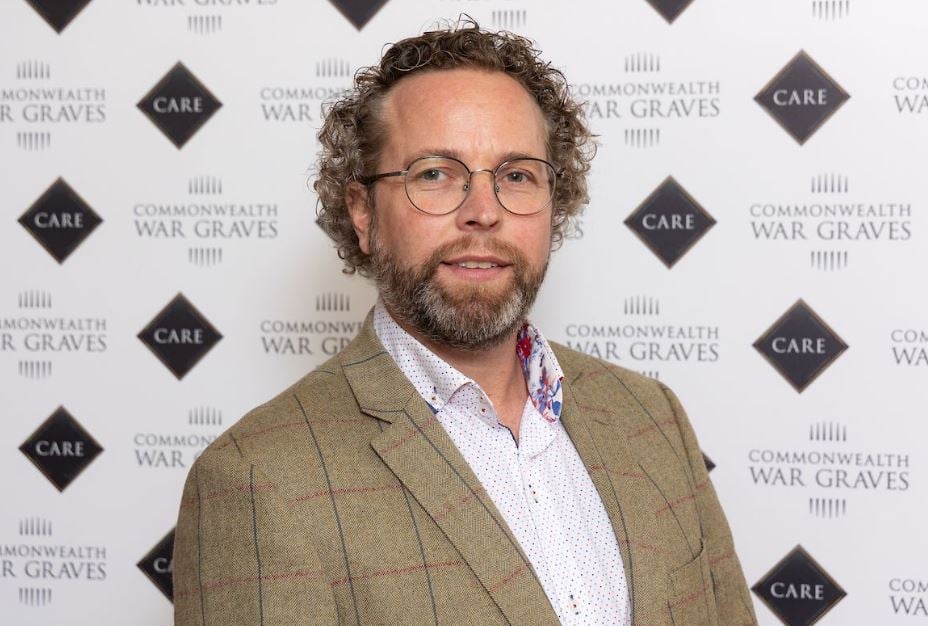 Man with curly hair, beard, and glasses wears a tweed jacket and patterned shirt. Background displays "Commonwealth War Graves" logo.