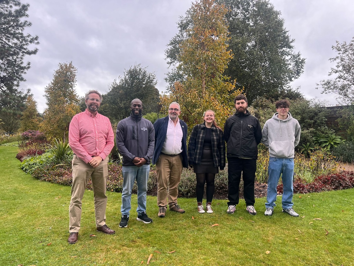 Six people pose for a photo in an outdoor garden setting, with trees and autumnal foliage in the background.