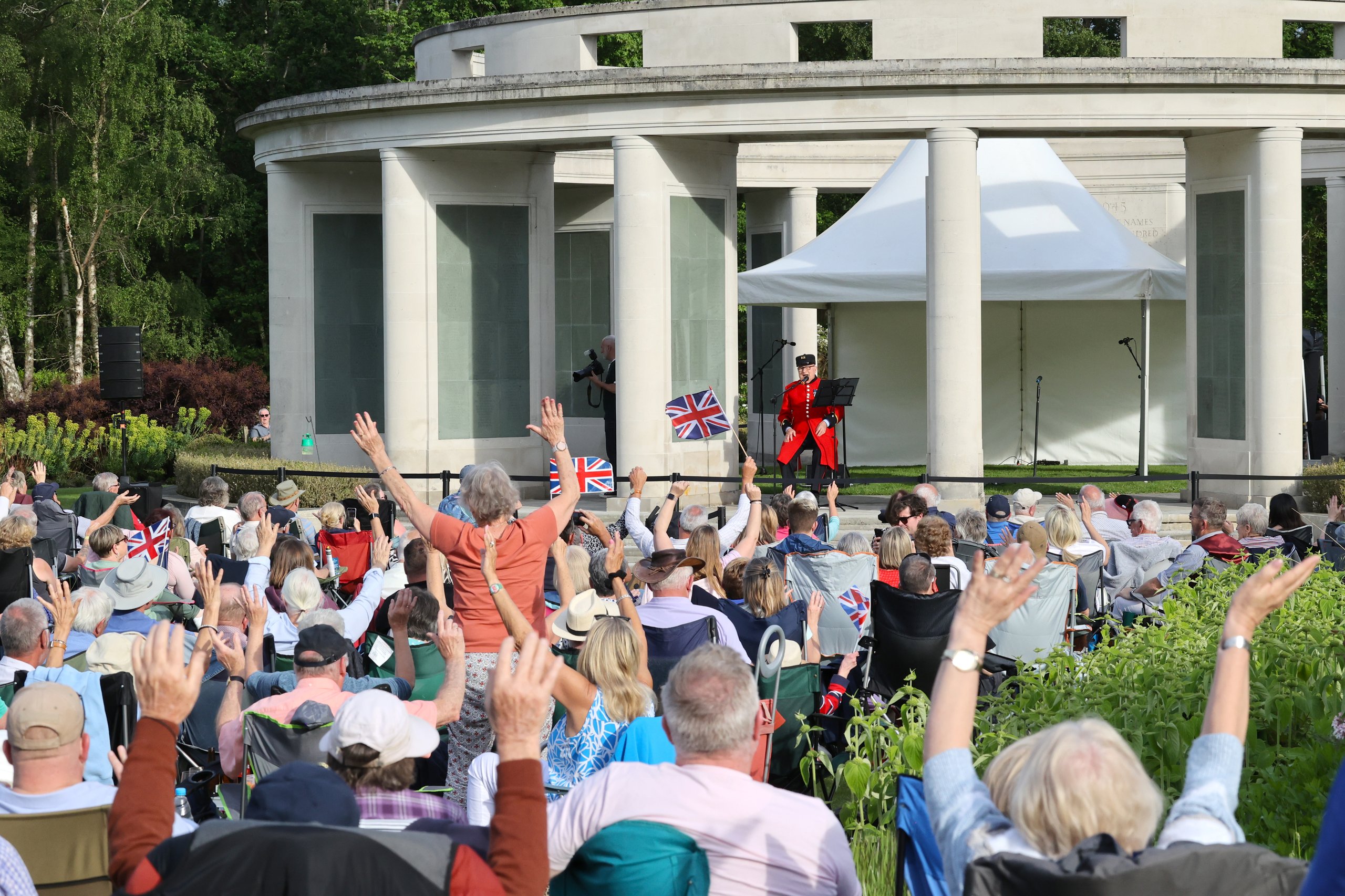 An audience watches a ceremony with a person in a red uniform speaking. Many attendees hold up their hands and Union Jack flags.