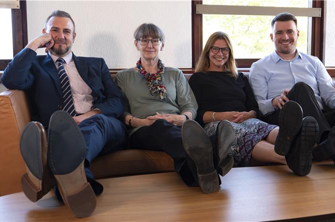 Four casually dressed people sit on a sofa with their feet up on a coffee table.