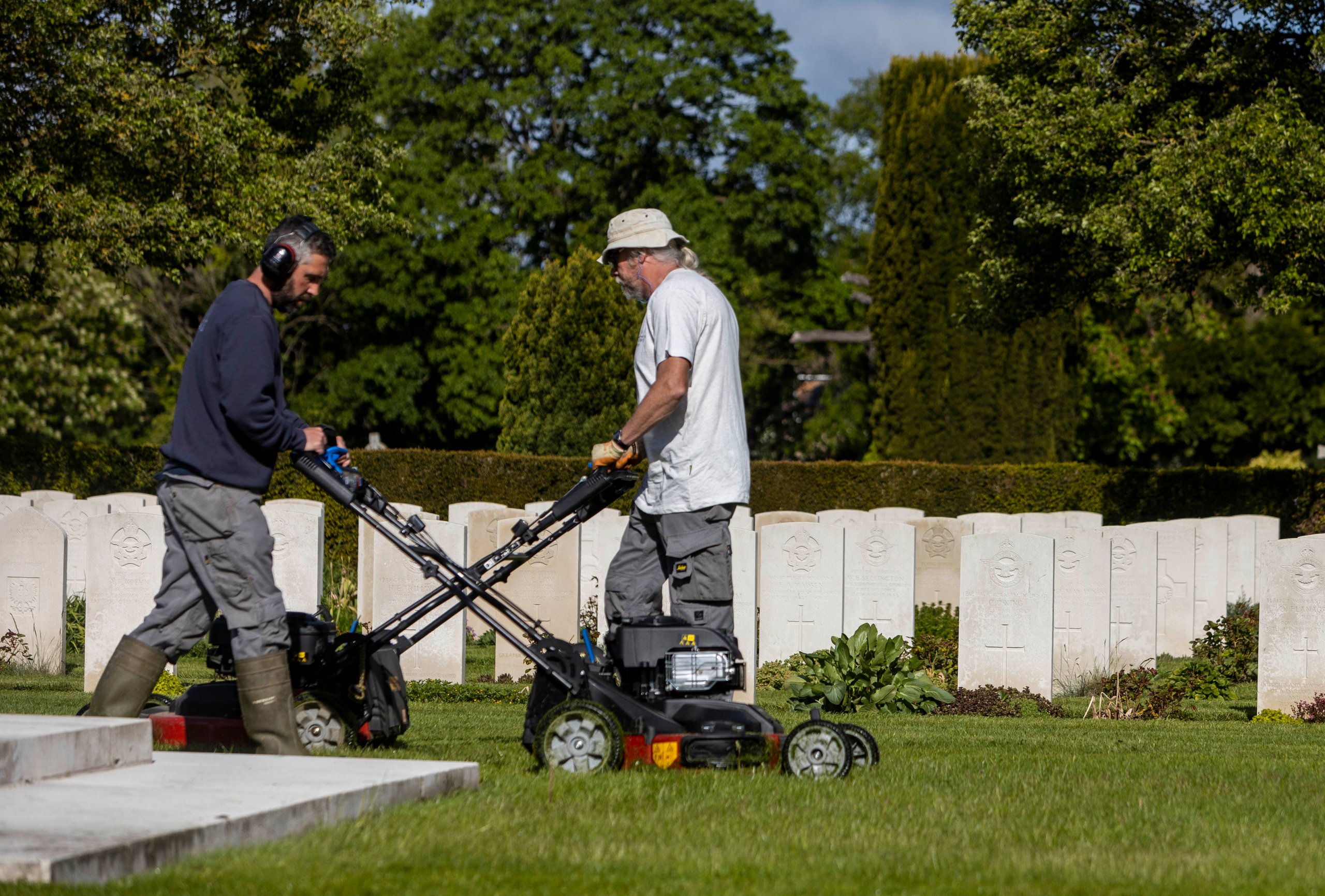 Two men with lawnmowers tend to the grounds of a cemetery with rows of white headstones and trees.