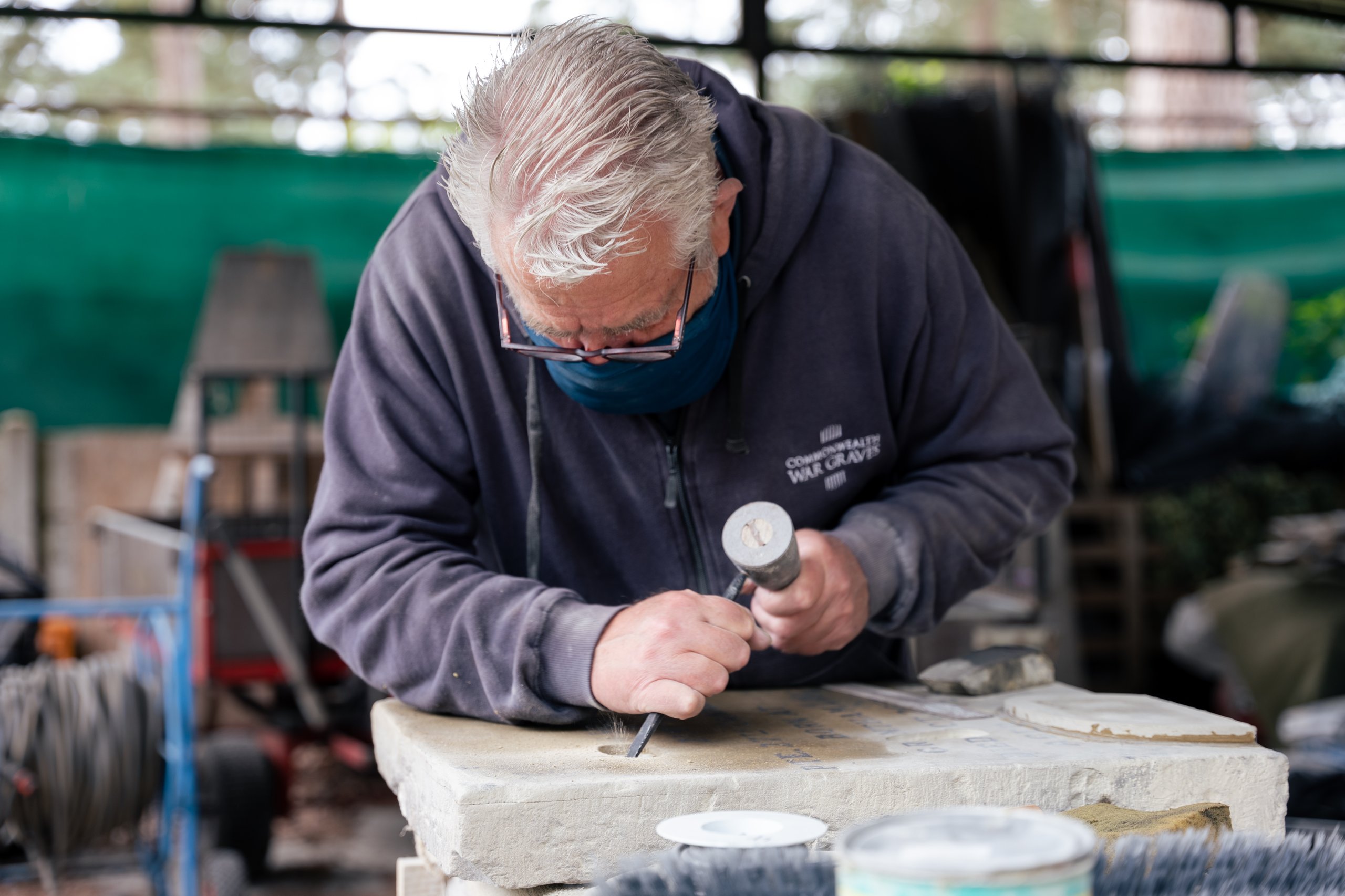 A man in a dark blue hoodie and mask carves into a stone slab with a chisel and mallet in a workshop.