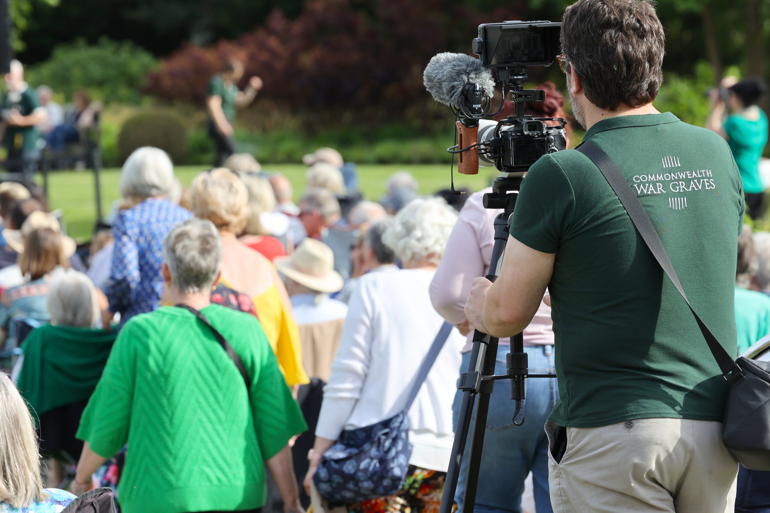 A cameraman films an outdoor event with attendees in the background. The cameraman wears a green polo shirt with "Commonwealth War Graves" printed on it.