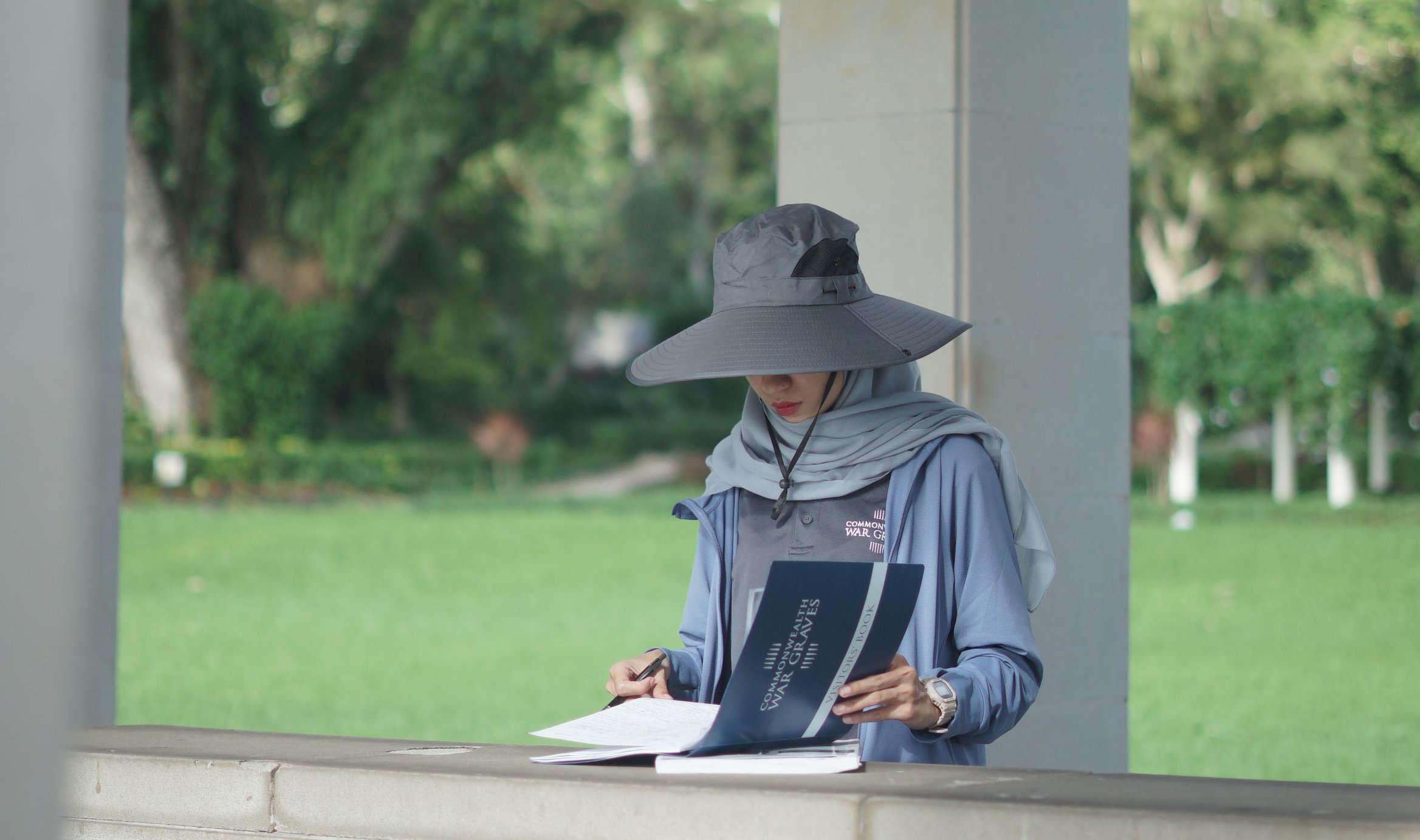 A person wearing a sun hat and hijab reads a visitor book at a war memorial, with trees and grass in the background.